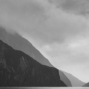 Black and white fine art photography print of dramatic Milford Sound cliffs and misty peaks with a tiny cruise ship revealing the vast scale