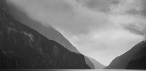 Black and white fine art photography print of dramatic Milford Sound cliffs and misty peaks with a tiny cruise ship revealing the vast scale
