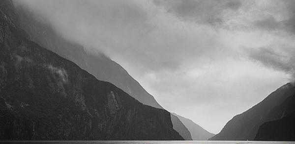 Black and white fine art photography print of dramatic Milford Sound cliffs and misty peaks with a tiny cruise ship revealing the vast scale